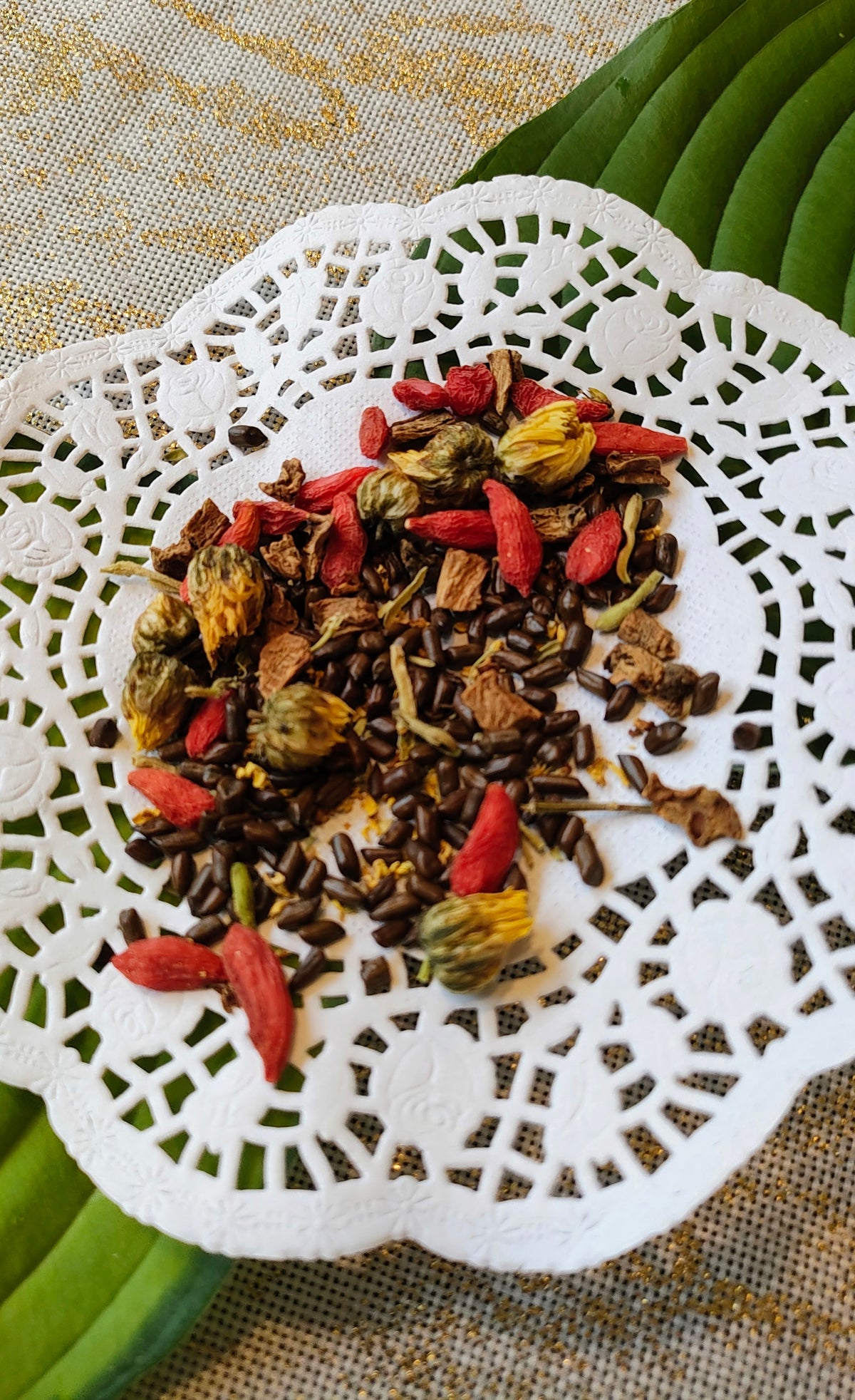 A close-up of loose herbal tea ingredients, including dark brown seeds, dried red berries, and yellow flowers, arranged on a white lace doily over a textured beige fabric with green leaves in the background.