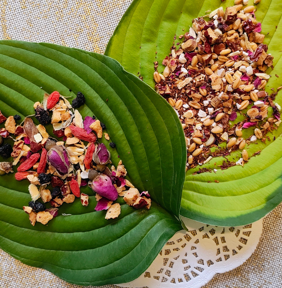 A mix with dried rose petals, seeds, and berries presented on large green leaves over a woven beige fabric background.