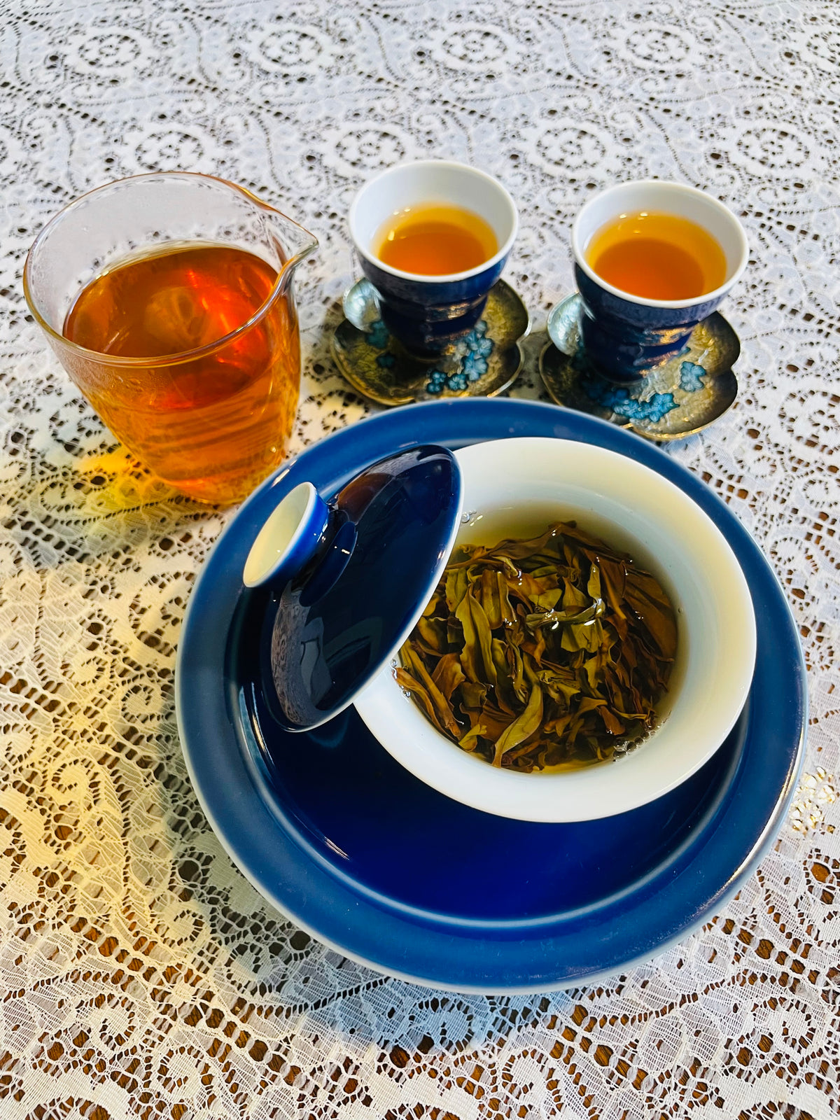 A tea set arranged on a white lace tablecloth, featuring a blue and white gaiwan with a lid, two matching teacups with saucers, and a glass pitcher filled with amber-colored tea.