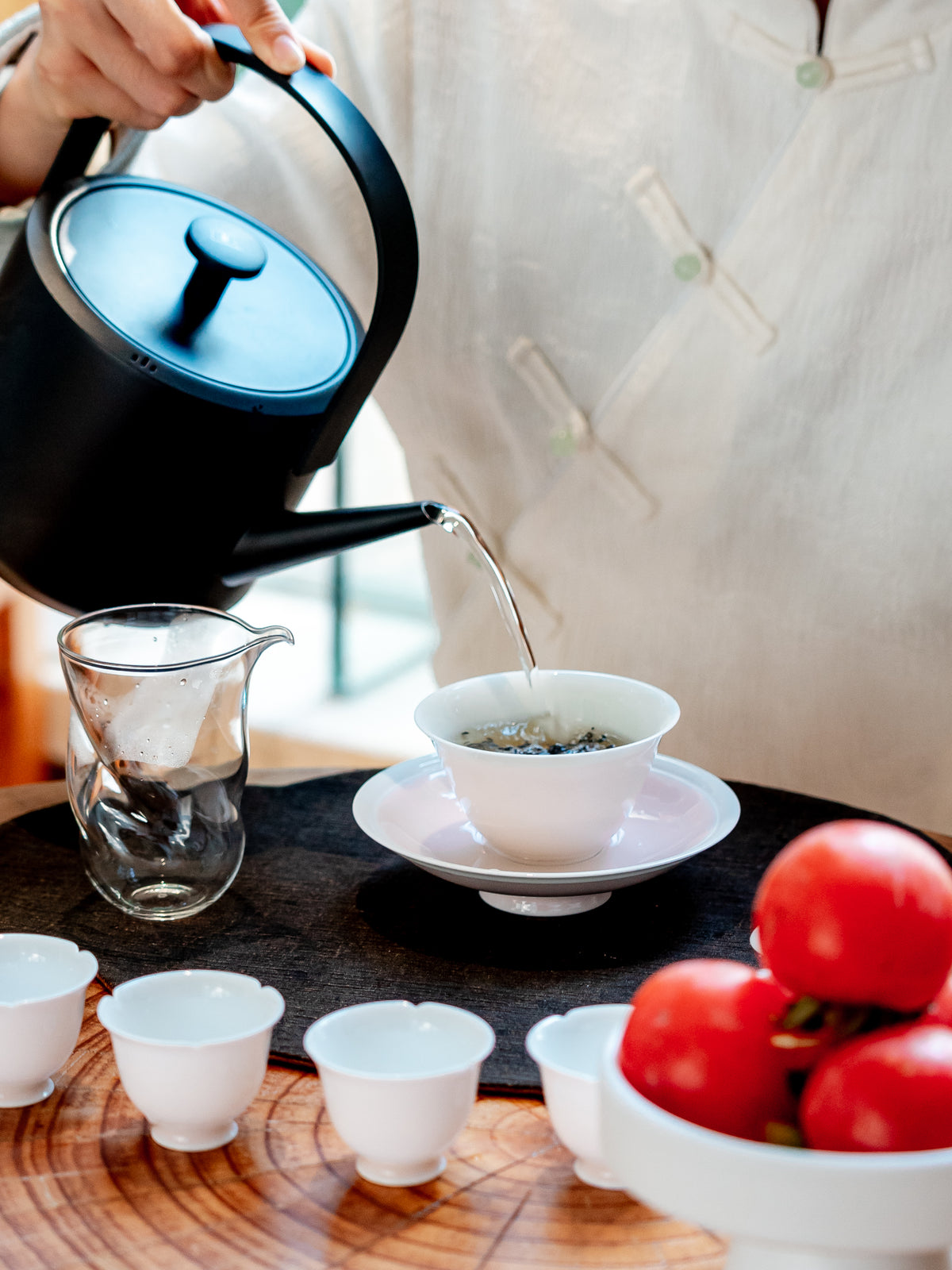 A person in a white traditional Chinese top pours hot water from a black teapot into a white gaiwan bowl on a saucer, with small white tea cups and red tomatoes arranged on a wooden table.