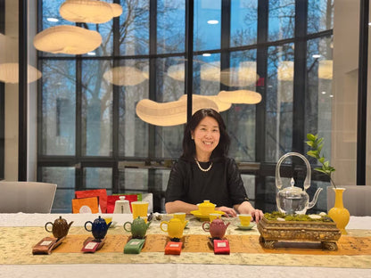 A woman sits behind a long table displaying a collection of colorful teapots, cups, and a glass teapot in a modern interior with large windows and hanging pendant lights.