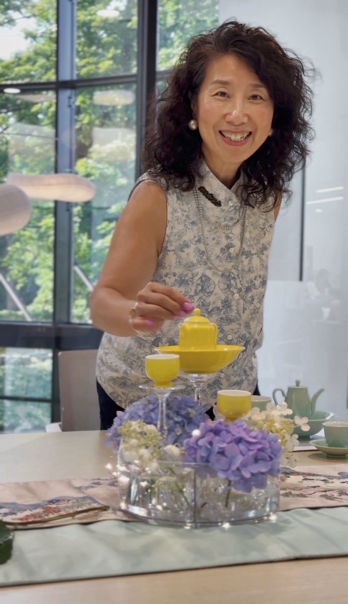 A smiling woman with curly dark hair stands behind a table, holding a small yellow teapot above matching yellow cups. The table is decorated with purple hydrangeas in a clear glass tray and a teapot, set in a bright room with large windows and greenery outside.