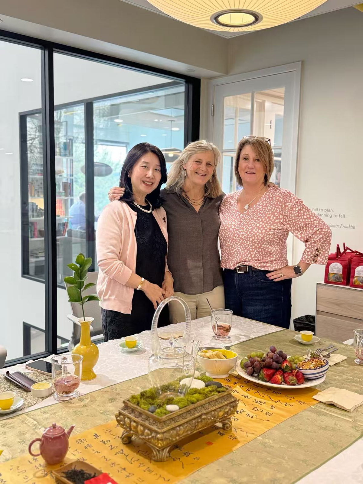 Three women posing together in a bright, modern room with large windows, standing behind a decorated table with tea sets, fruit, and a decorative tray.