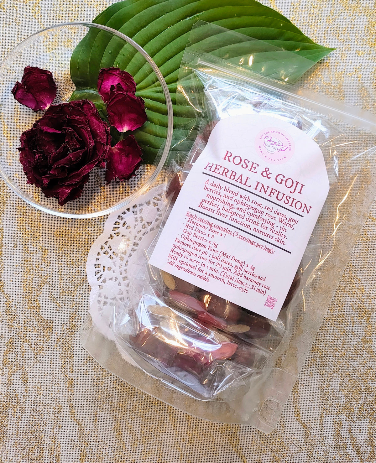 A clear plastic bag labeled 'Rose & Goji Herbal Infusion' containing dried rose petals and goji berries, styled on a beige patterned fabric with a green leaf, a glass bowl of dark red rose petals, and a white lace doily.