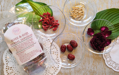 A clear plastic bag labeled 'Rose & Goji Herbal Infusion' sits beside several glass bowls containing dried red goji berries, beige seeds, red jujubes, and dark purple rose petals on a patterned beige surface.