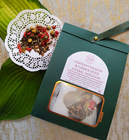 A dark green tea pouch labeled 'Chrysanthemum & Cassia Seed Herbal Infusion' with a clear window showing tea bags inside, placed on a textured beige surface beside a white lace doily holding loose herbal tea ingredients. A large green leaf is visible on the left.