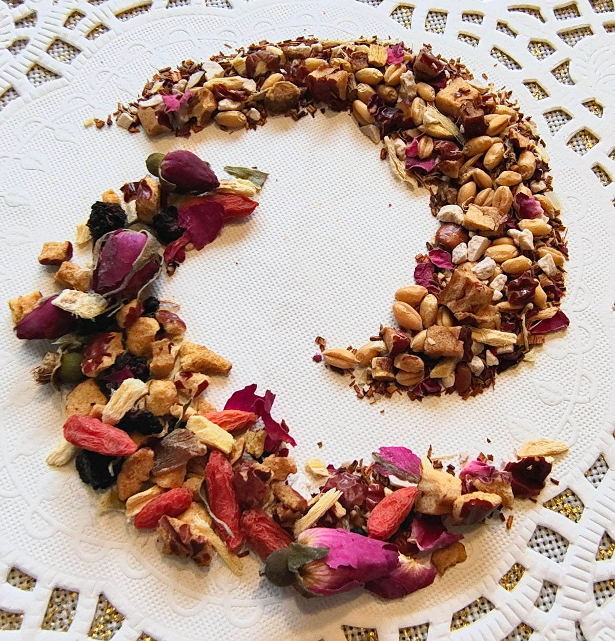 A circular arrangement of loose-leaf herbal tea or spice blend made from almonds, rose petals, dried berries, and other botanicals, presented on a white textured plate.