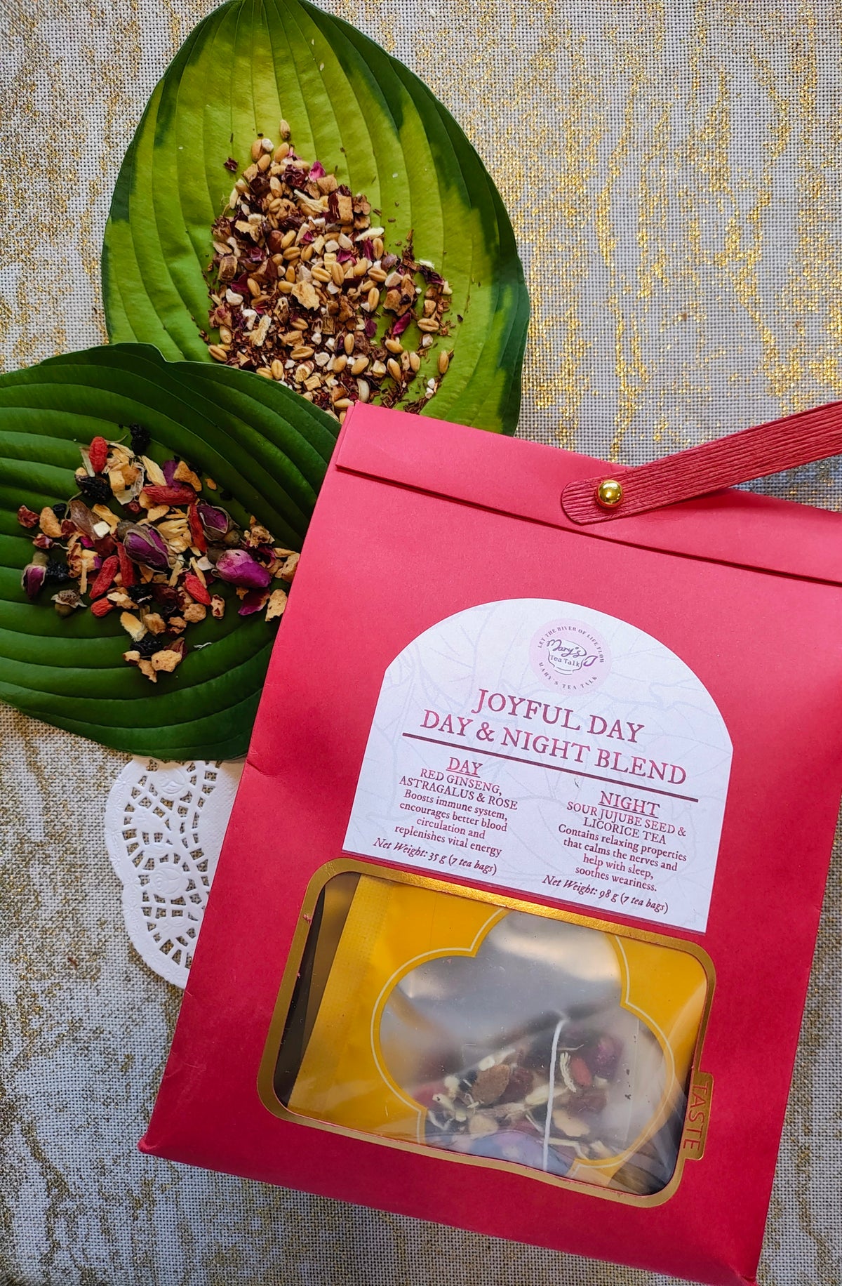 A red paper bag of 'Joyful Day & Night Blend' herbal tea with a clear window showing the tea inside, styled on a textured gold background with green leaves holding loose tea blends.