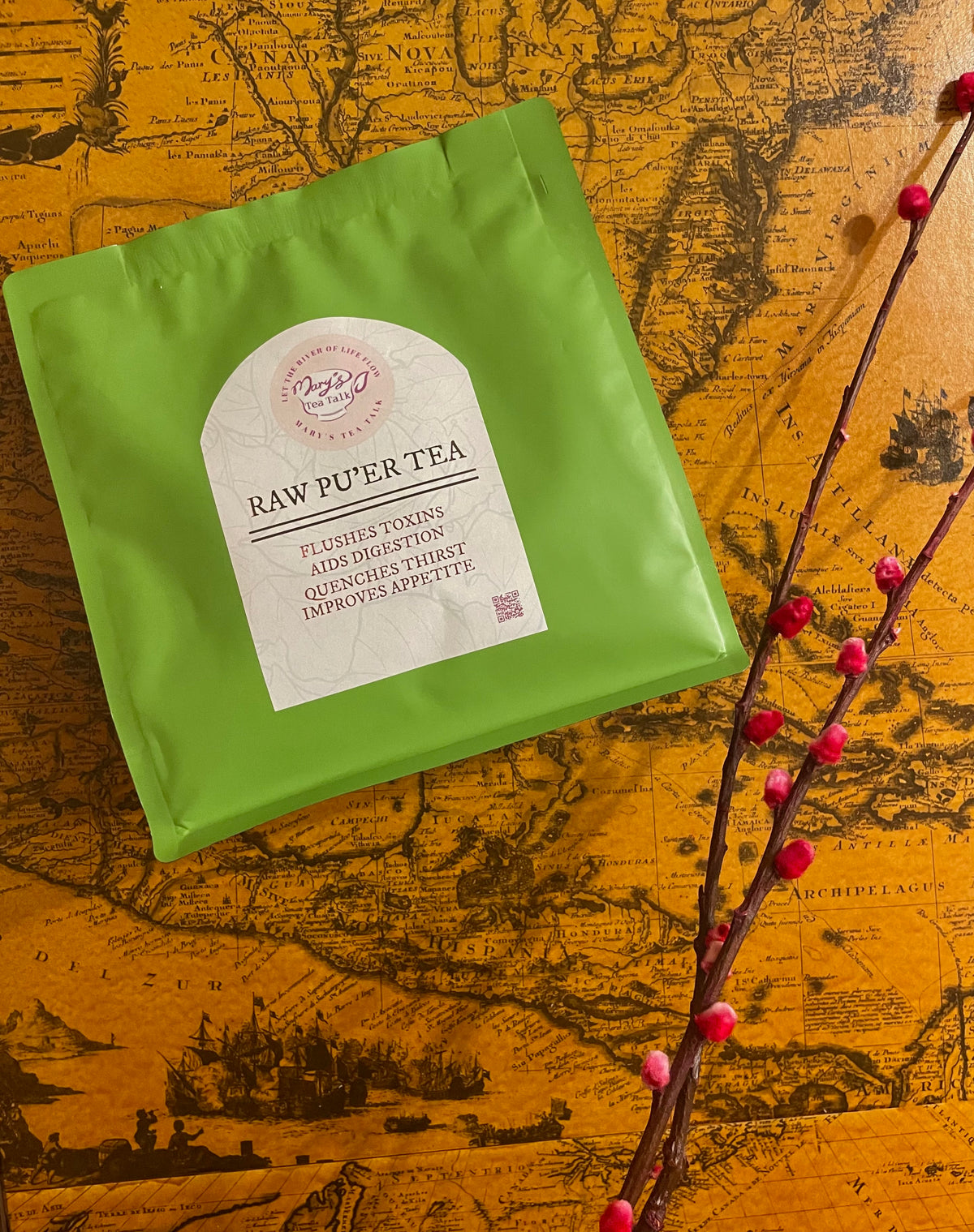A green tea bag pouch labeled 'RAW PU'ER TEA' placed on an antique-style map, with a decorative branch of red berries running diagonally across the right side.