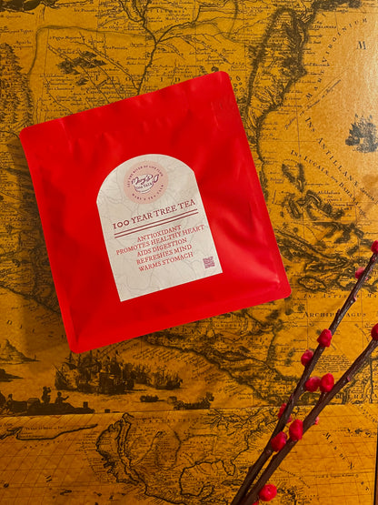 A red pouch of 100 Year Tree Tea with a white label, placed on a vintage-style map background with decorative red berries in the foreground.
