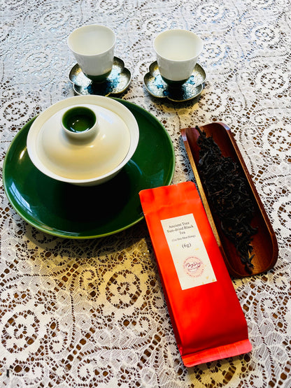 A tea set arrangement on a white lace tablecloth, featuring a green lidded teapot bowl, two white cups with saucers, a red-orange pouch labeled 'Natural Black Tea', and a dark tray of loose tea leaves.