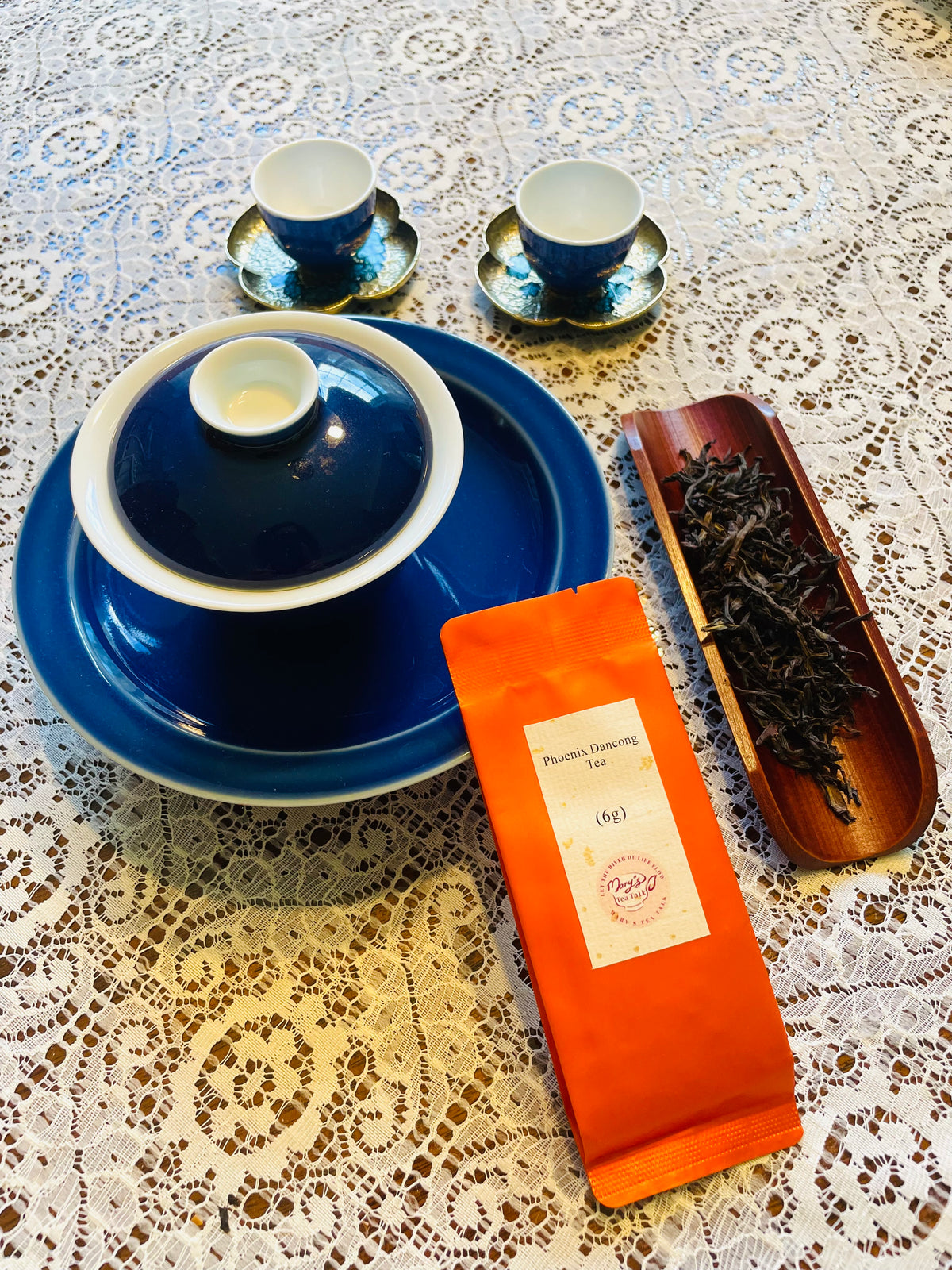 A tea set arrangement on a white lace tablecloth, featuring a dark blue ceramic gaiwan with lid, two matching blue teacups and saucers, an orange packet labeled Phoenix Dancong Tea, and a wooden tray holding loose dried tea leaves.
