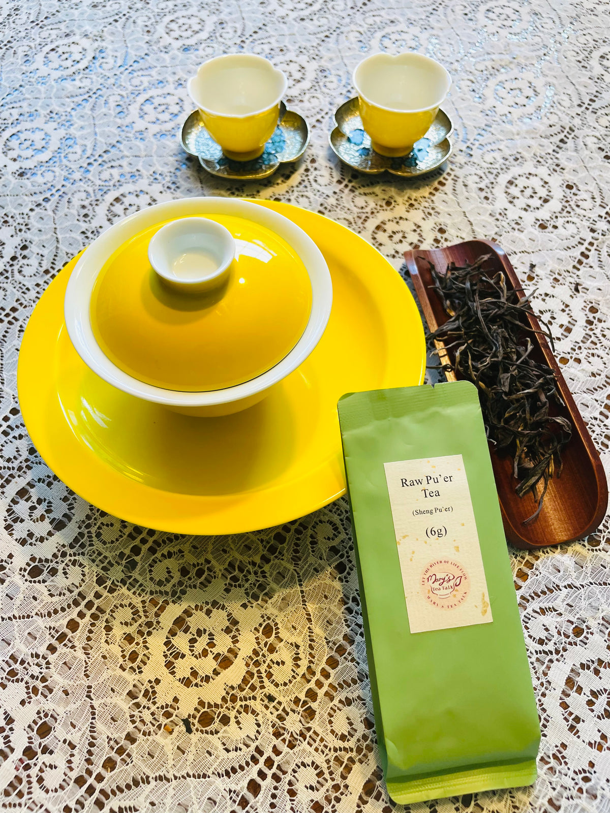 A tea set arrangement on a lace tablecloth, featuring a bright yellow lidded teapot on a matching yellow plate, two small yellow teacups on saucers, a green pouch of raw pu'er tea, and a wooden tray holding loose dried tea leaves.