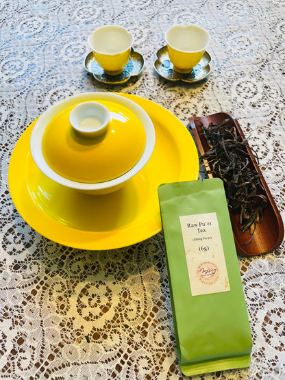 A tea set arrangement on a lace tablecloth, featuring a bright yellow lidded teapot on a matching yellow plate, two small yellow teacups on saucers, a green pouch of raw pu'er tea, and a wooden tray holding loose dried tea leaves.