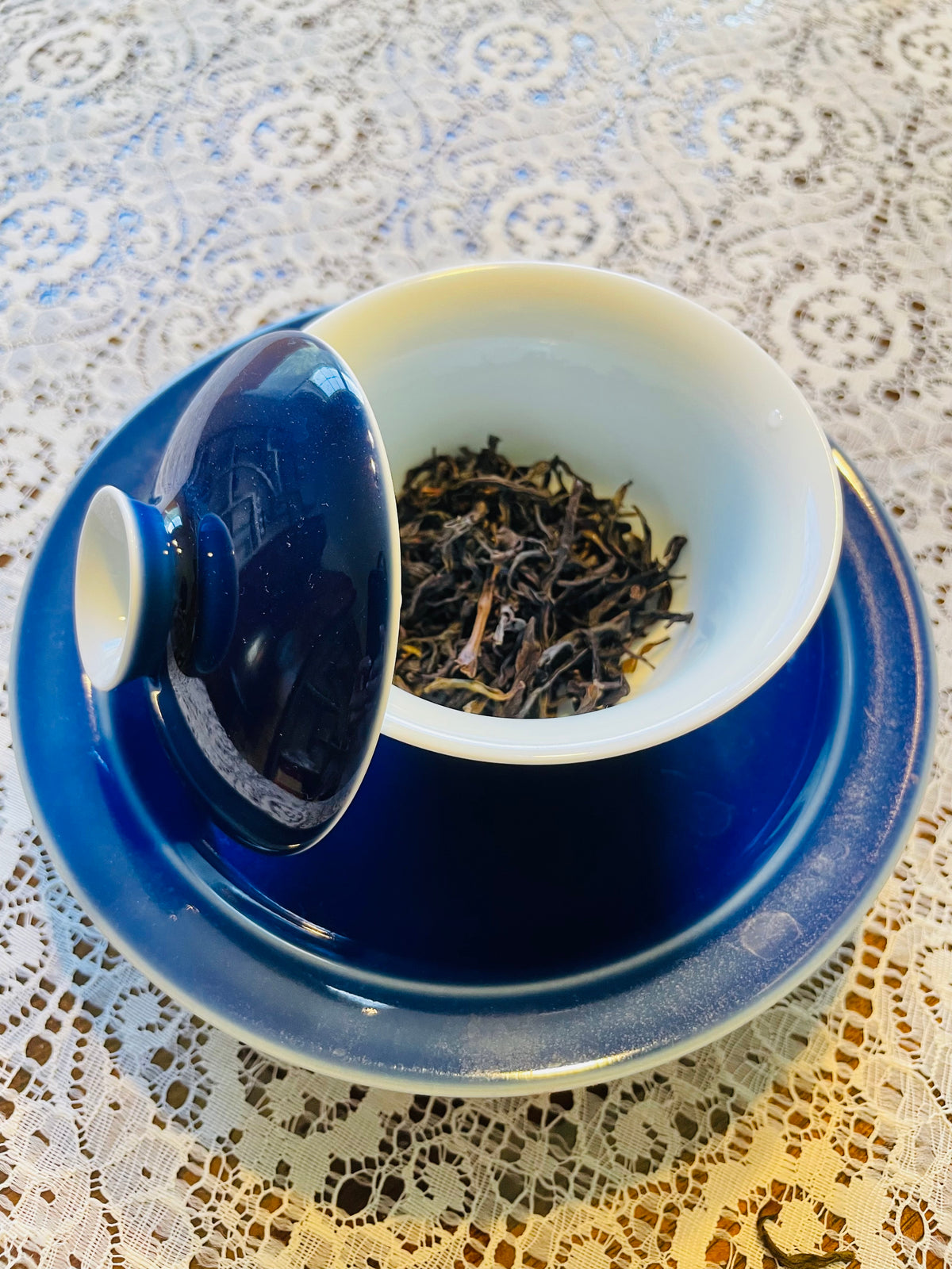 A cobalt blue and white ceramic gaiwan set with a dark loose-leaf tea inside, placed on a white lace tablecloth.