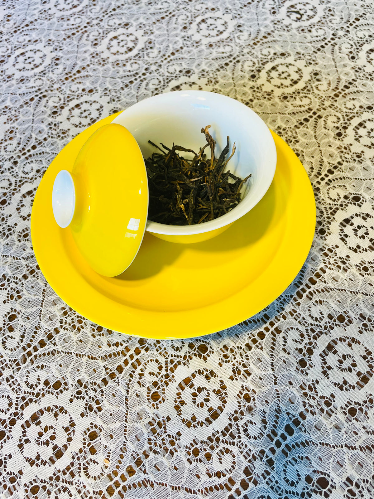 A yellow and white ceramic tea set with a matching lid, containing loose-leaf tea, placed on a yellow saucer over a white lace tablecloth.