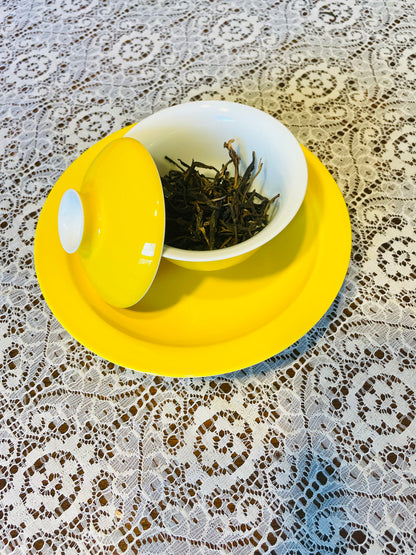 A yellow and white ceramic tea set with a matching lid, containing loose-leaf tea, placed on a yellow saucer over a white lace tablecloth.