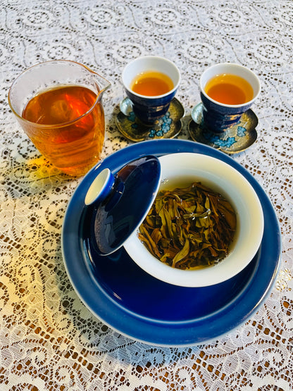 A tea set arranged on a white lace tablecloth, featuring a blue and white gaiwan with a lid, two matching teacups with saucers, and a glass pitcher filled with amber-colored tea.
