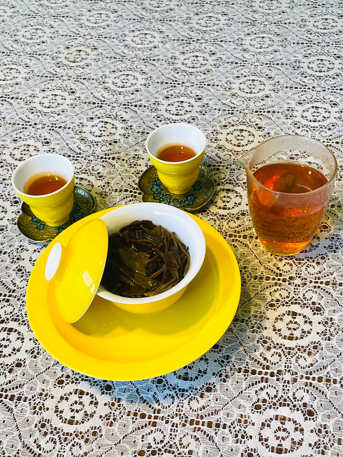 A yellow ceramic tea set arranged on a white lace tablecloth, including a yellow lidded bowl with dark tea leaves, two small yellow cups with amber tea, and a clear glass pitcher filled with tea.