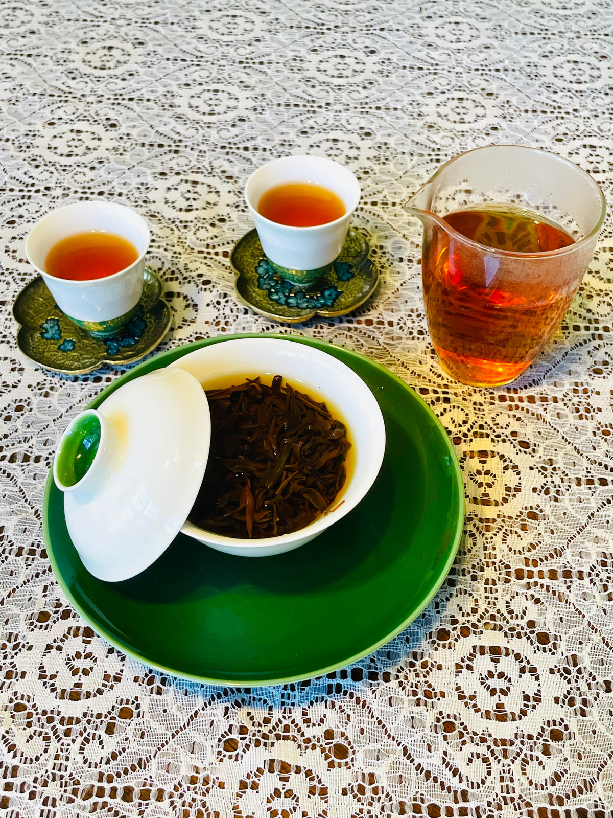 A tea set arranged on a white lace tablecloth, featuring a green plate with a white gaiwan bowl containing loose tea leaves, two small white cups filled with amber tea on decorative green and blue saucers, and a clear glass pitcher with more tea.