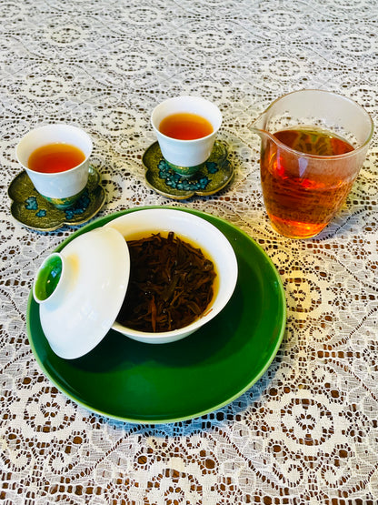 A tea set arranged on a white lace tablecloth, featuring a green plate with a white gaiwan bowl containing loose tea leaves, two small white cups filled with amber tea on decorative green and blue saucers, and a clear glass pitcher with more tea.