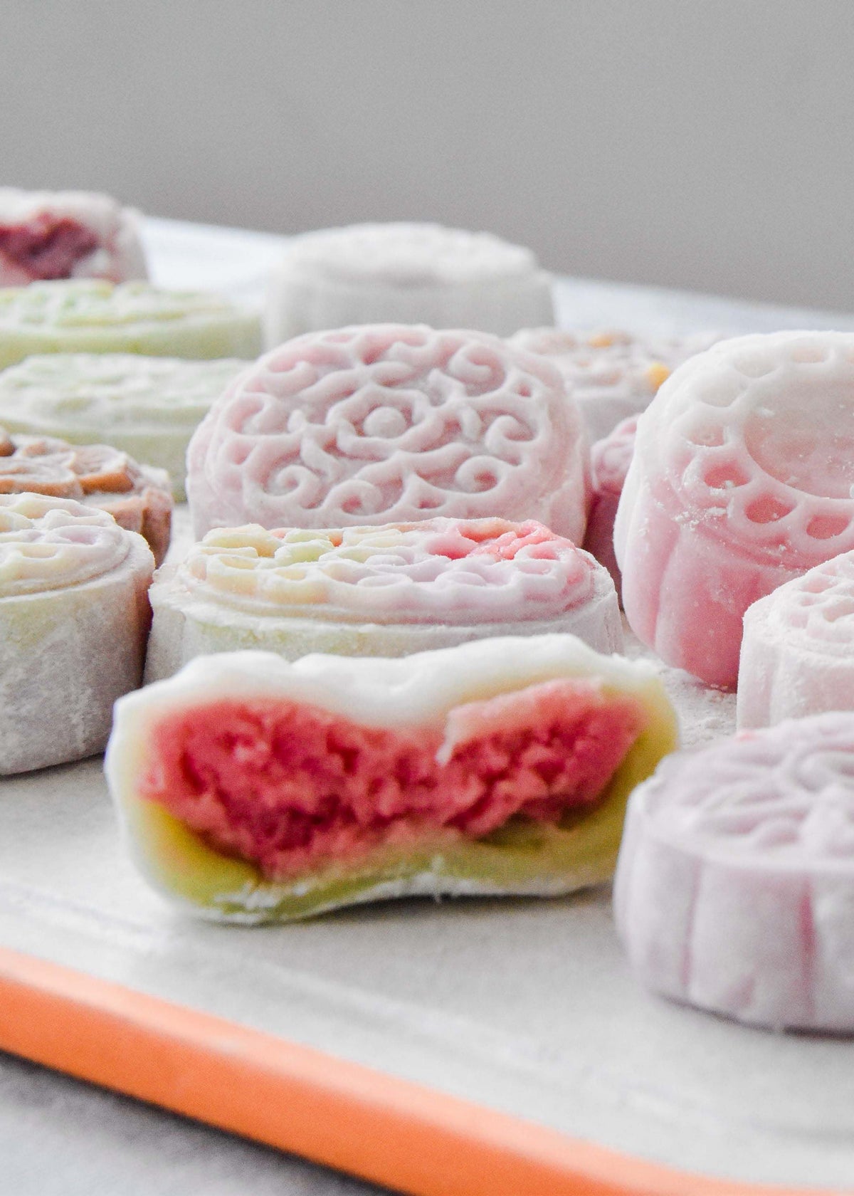 A close-up of assorted mooncakes arranged on a tray with an orange edge. Several round pastries are dusted with white powder, while one cut-open pastry in the foreground reveals pink red filling and a pale green layer.