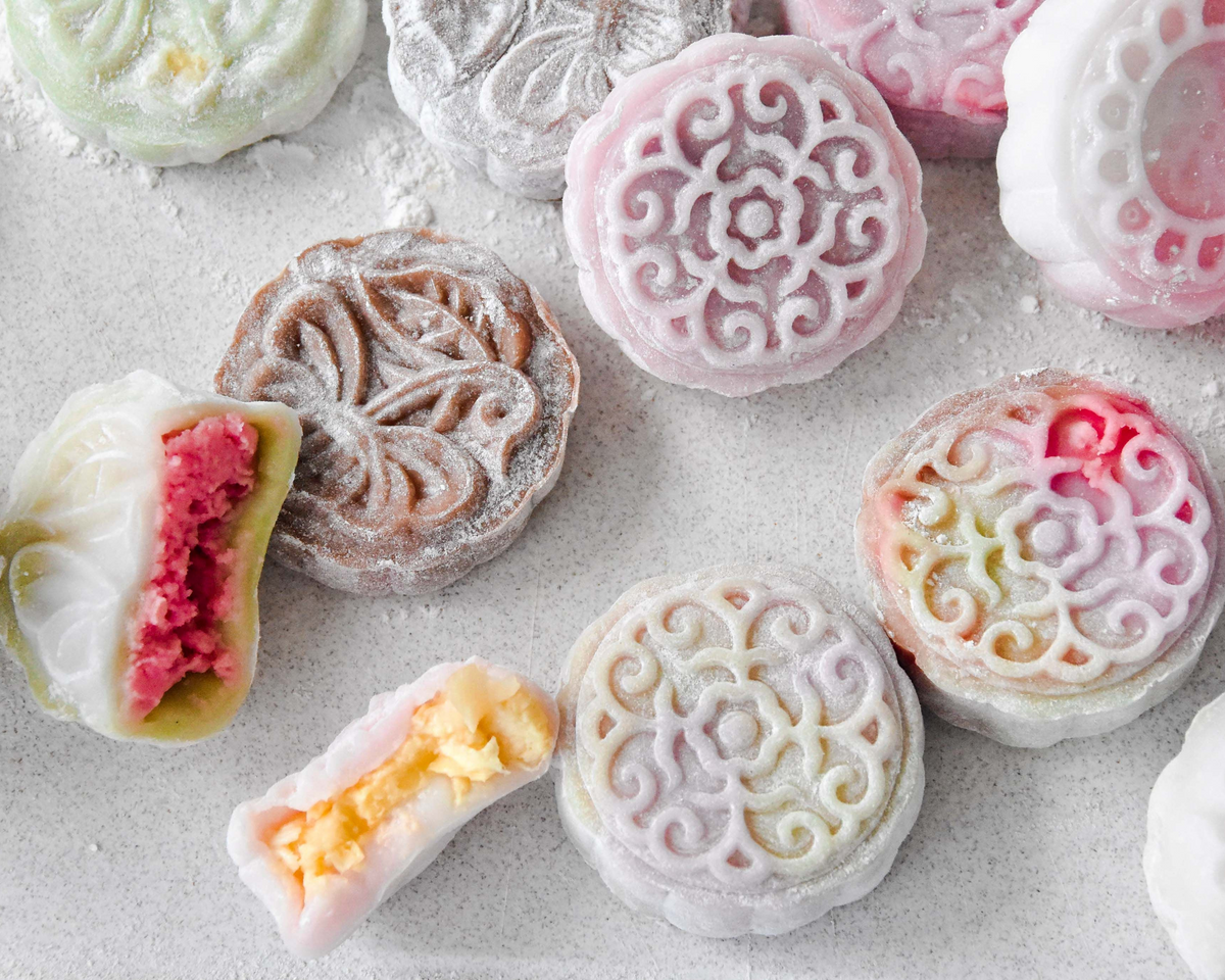 A close-up flat lay of colorful moon cakes on a light gray surface, with some cut in half to show pink and yellow fillings. The cakes have intricate floral and scroll patterns in shades of pink, white, brown, green, and yellow.