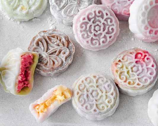 A close-up flat lay of colorful moon cakes on a light gray surface, with some cut in half to show pink and yellow fillings. The cakes have intricate floral and scroll patterns in shades of pink, white, brown, green, and yellow.