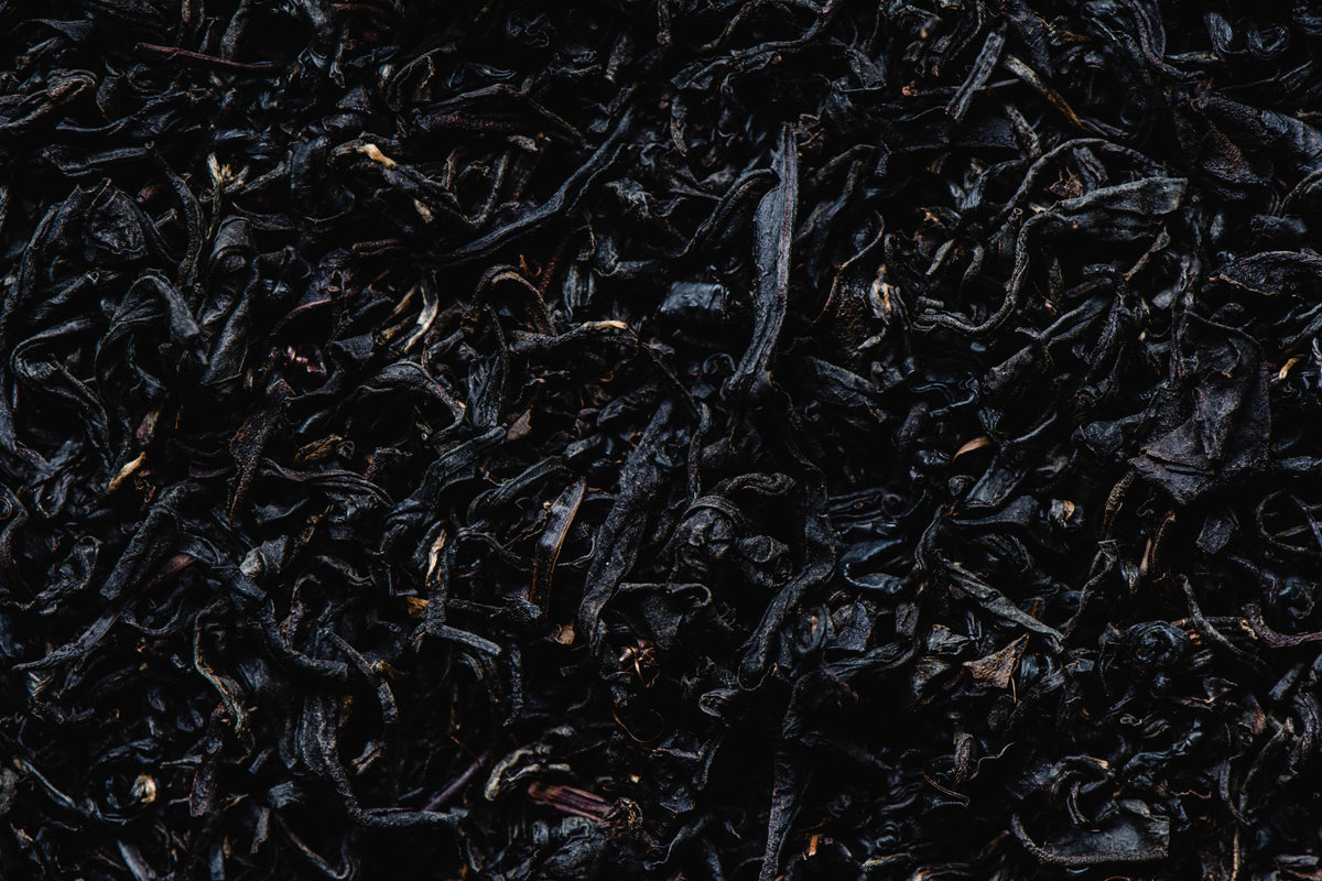A close-up texture shot of dark, loose-leaf tea leaves, appearing very dark brown to black with subtle reddish-brown and golden-brown highlights.